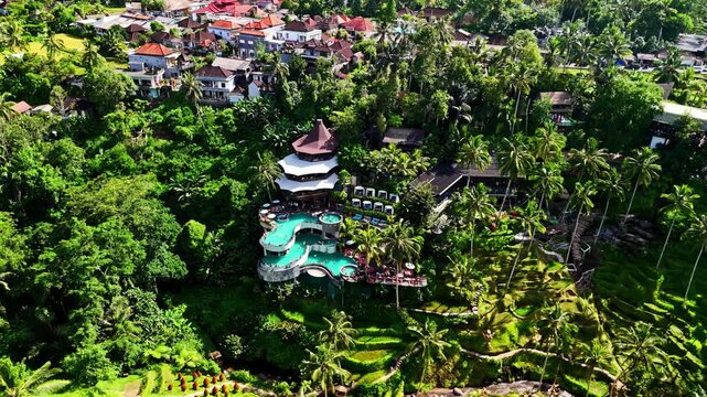 Aerial view of a luxury resort with tiered infinity pools nestled in a lush tropical jungle surrounded by palm trees and nearby village rooftops in Bali, Indonesia.