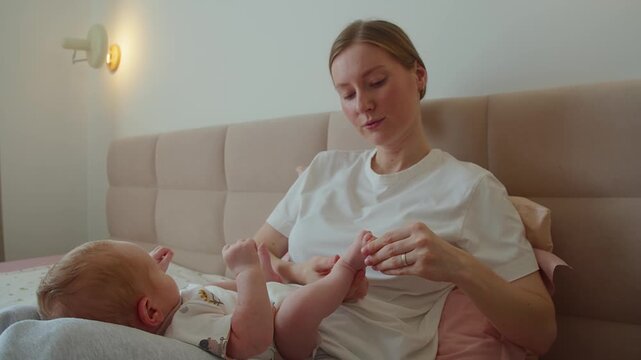 Mother talks to baby while massaging toes during quiet care on bed. Warm voice and gentle touch create comfort, sensory learning, trust, emotional connection, and attentive home caregiving.