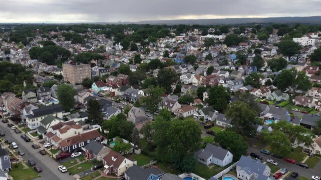 Dense residential rooftops stretch across cloudy suburban neighborhood in Belleville, New Jersey, cars travel curved street past backyard pools and tall apartment tower. Aerial drone flyover shot.