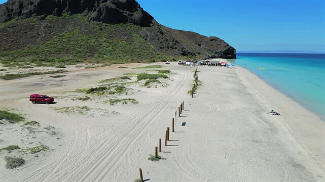 Drone flies low over El Tecolote Beach near Punta Shiro, following a line of wooden posts along the shore toward a group of tents at the end of the beach.