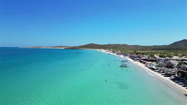 Drone flies in toward El Tecolote Beach near La Paz, passing a large beachfront restaurant with gathered crowds and swimmers speckled in the water, slowly pulling out toward the ocean as it passes.
