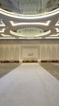 The calm before a high-profile Middle Eastern gala. A pristine white runner splits the patterned carpet of a grand venue glowing under massive crystal chandeliers.