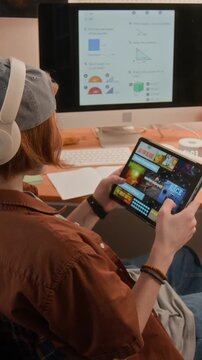 Vertical video of Caucasian teenager wearing headphones playing mobile game on tablet instead of studying math on computer. Ideal for e-learning and digital distraction concepts