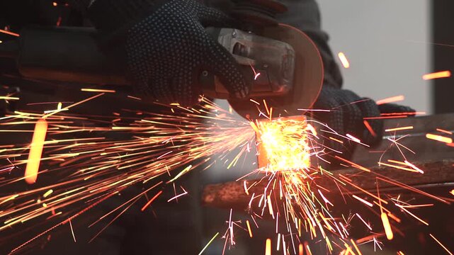 Gloved worker grinding square steel tube with handheld angle grinder, sending bright orange sparks through dim industrial workshop, rough metal textures, dark workwear, blurred gray background