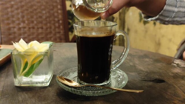 Serving signature Robusta black coffee from Java, Indonesia. a shot showing sugar syrup being added to a glass of coffee. The setting is a quiet, warm room perfect for relaxing and enjoying the coffee