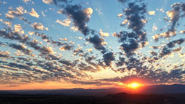 4K drone timelapse of a vibrant sunrise bursting over a rugged mountain and canyon landscape. Distinct rows of clouds move rhythmically as radiant sunbeams project upward into the blue dawn sky.