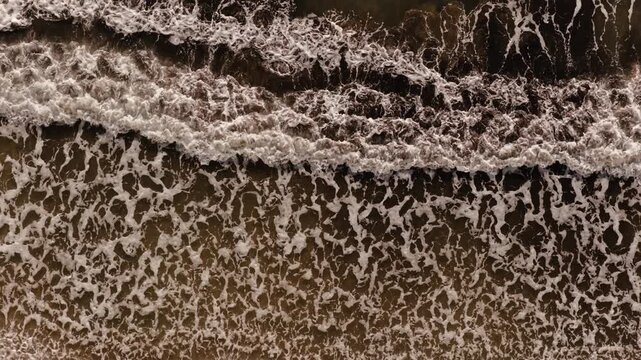 Overhead drone view of foamy ocean waves washing onto a sandy beach on Corfu's coastline.