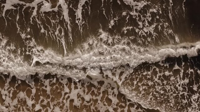 Overhead drone close-up of a breaking wave with intricate sea foam patterns along Corfu's sandy shoreline.