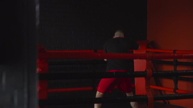 Focused boxer standing inside dim boxing ring with red ropes, holding yellow training wraps near face while keeping guarded stance beside bright corner padding, with boxing, boxer, training, fighter