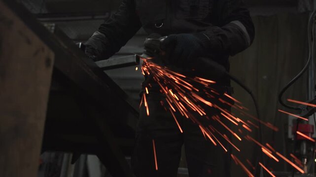 Worker using handheld grinder on metal inside dim industrial workshop, sending bright orange sparks across frame beside dark protective workwear, cables, steel surfaces, and gritty machinery