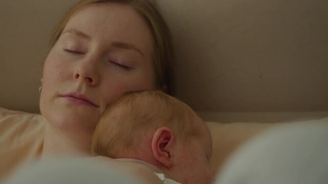 Close-up shows young mother asleep while holding baby securely in her arms. Relaxed face and protective posture coexist, suggesting exhaustion, trust, shared warmth, and brief maternal recovery.