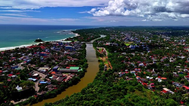 Aerial view of a river flowing into the Indian Ocean through a coastal town with lush tropical vegetation and sandy beaches under a cloudy blue sky in Sri Lanka.
