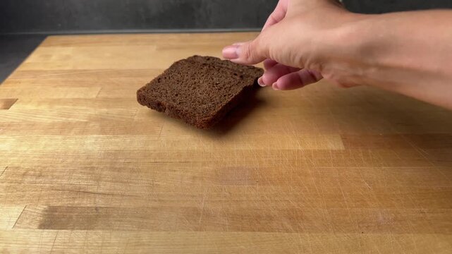 A woman's hand places slices of dark rye bread onto a wooden cutting board in a domestic kitchen setting