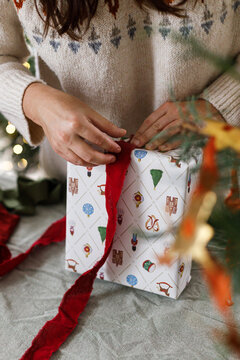Female hands wrapping winter holiday present with stylish paper and red bow. Cozy Christmas eve atmosphere, handmade gift preparation, scandinavian style interior decoration.