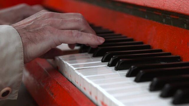 Adult hands pressing worn keys on red upright piano in tight horizontal close-up with visible finger contact, textured skin, key depression, and tactile performance detail