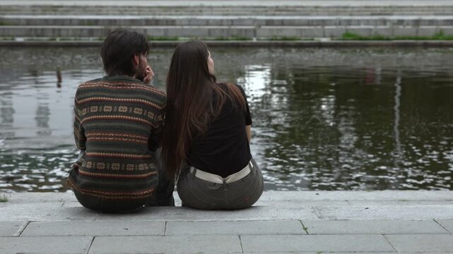 Two young adults seated on stone steps beside urban pond, one pointing across water while turning toward companion, quiet outdoor moment with relaxed body language