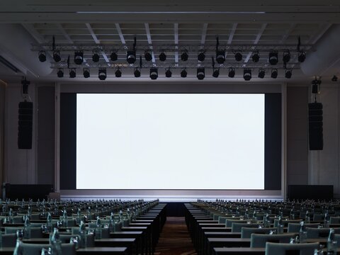 Blank white screen mockup on stage in conference hall seminar room with Rows of empty seats and lighting setup