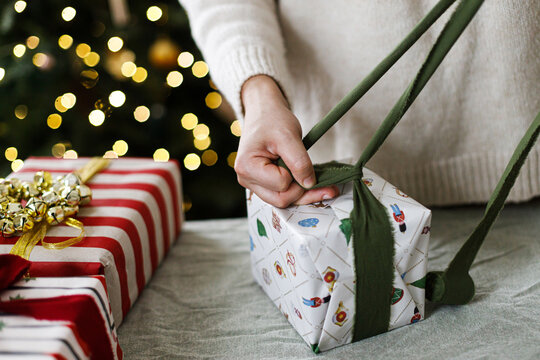 Female hands wrapping winter holiday present with stylish paper and green bow. Cozy Christmas eve atmosphere, handmade gift preparation, scandinavian style interior decoration.
