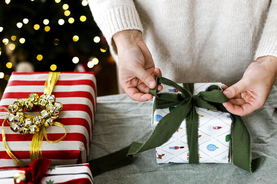 Wrapping stylish xmas presents. Woman in cozy sweater wrapping Christmas gift box with green ribbon at home. Festive DIY holiday preparation near decorated xmas tree with golden bokeh lights