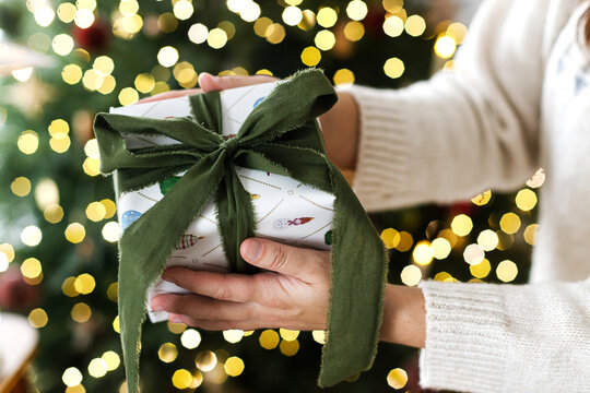 Merry Christmas! Woman in warm knitted sweater holding beautifully wrapped Christmas gift box with green ribbon. Close up of hands giving xmas present, festive tree lights on background