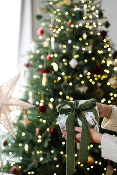 Merry Christmas! Woman in warm knitted sweater holding beautifully wrapped Christmas gift box with green ribbon. Close up of hands giving xmas present, festive tree lights on background