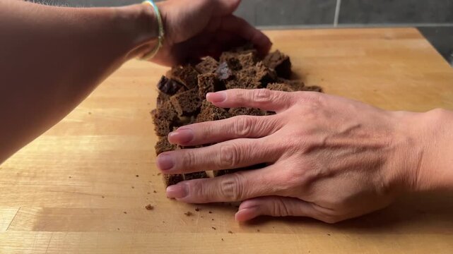 A person's hands gather a pile of diced dark rye bread cubes together on a wooden cutting board in a home kitchen