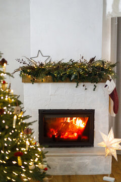 Warm fire burning in fireplace with Christmas decorations on mantelpiece. Cozy scandinavian living room interior on xmas eve, traditional winter holidays atmosphere at home.