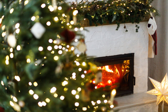 Cozy burning fireplace with warm flame decorated with pine garland, stocking and paper star lamp. Decorated Christmas tree with glowing lights on background, festive winter interior