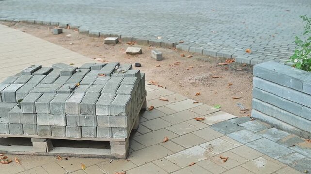 A stack of gray bricks sits on a wooden pallet on a sidewalk next to a paved area. The setting shows an outdoor construction site with some scattered materials and soil around