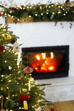 Christmas tree with beautiful baubles, garland lights and paper ornaments. Warm fire burning in cozy fireplace on background. Magical xmas eve atmosphere, traditional home decoration.