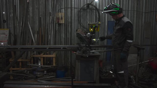 Helmeted metalworker handling material beside cutting saw in dim industrial workshop with corrugated wall, cables, stacked bars, cluttered tools, and moody overhead light
