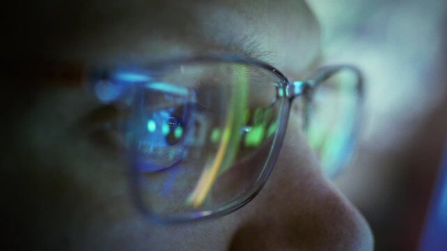 Bespectacled system administrator sits in a server room at night, monitoring equipment to maintain system stability. The indicator light reflects in his glasses, underscoring reliability and control
