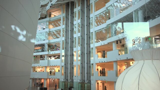 Multi level atrium interior with three glass elevator shafts, open balconies, clear railings, mixed daylight and warm ambient lighting across modern office lobby space