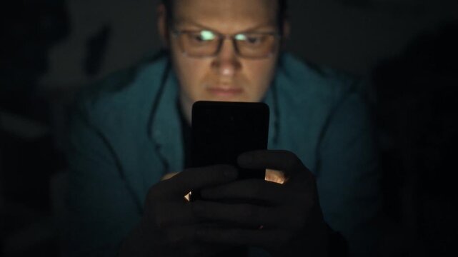 Man sits in darkness, eyes glued to glowing smartphone screen as he scrolls, embodying nightly digital addiction, the bright gadget disrupts sleep, highlighting technology's intrusive impact on rest
