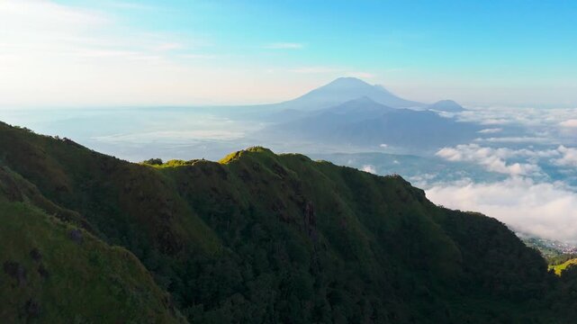 Aerial view of the lush green slopes of Mount Ungaran with clouds and distant peaks in Ungaran Barat, Central Java, Indonesia.