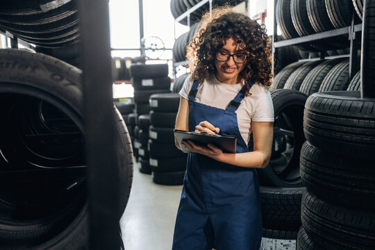 Female auto technician with curly hair wearing blue overalls checks inventory on a tablet surrounded by stacks of tires in a tire shop