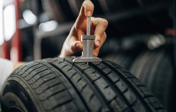 Hand holding a tire tread depth gauge measuring the tread on a car tire in a workshop with blurred tires in the background