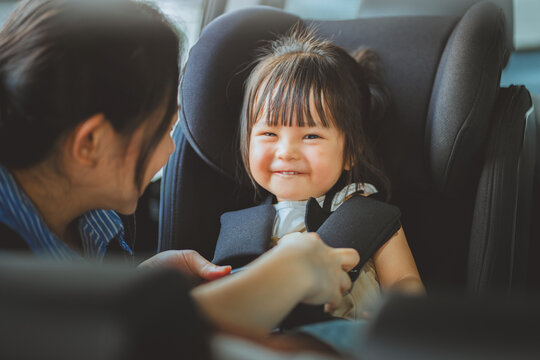 Asian mother fastening safety seat belt for cute smiling toddler girl in car seat before travel road trip, happy young mother securing child safety seat for family transportation, protection