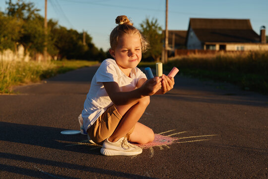A young girl sits on the ground in a quiet street, holding colorful chalk in her hands while she draws on the pavement during a sunny afternoon