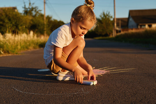 A child sits on the road and uses chalk to create drawings. It is evening, and sunlight casts a warm glow as grass and houses are seen behind