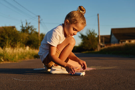 A young child sits on the road, focused on drawing with colorful chalk. The sun sets behind, casting a soft glow over the scene with houses in the background