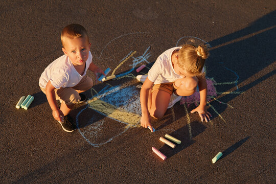 Two children focus on drawing with chalk on the pavement in a neighborhood during the late afternoon. Sunlight casts long shadows behind them