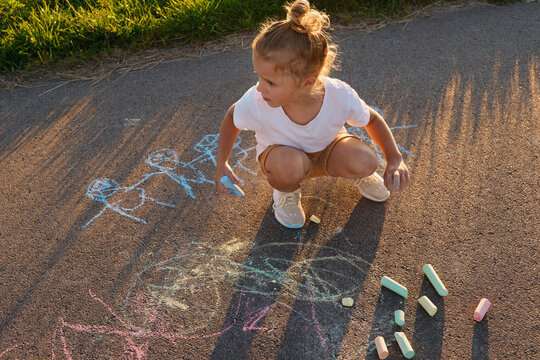 A child is sitting on the pavement using chalk to draw different pictures in the park at sunset. The child is focused on their artwork while enjoying the moment