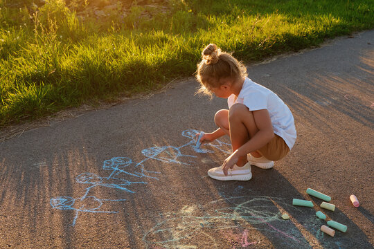 A child sits on a path writing with colorful chalk. The sun is low in the sky, casting shadows. Green grass surrounds the area. Chalk sticks are scattered nearby