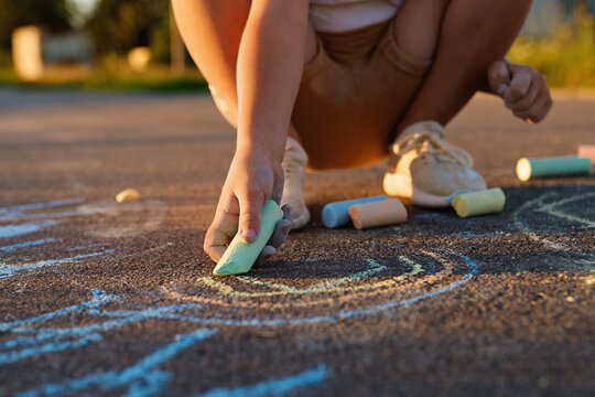 A child is crouching down to draw colorful shapes on the pavement using chalk. It is a warm evening in a park area with grass and trees nearby