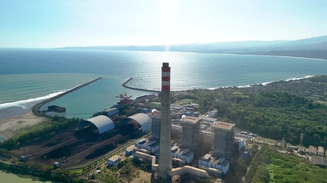 Aerial view of a coal-fired power plant with a tall chimney and industrial facilities situated along the coastline in Indonesia.