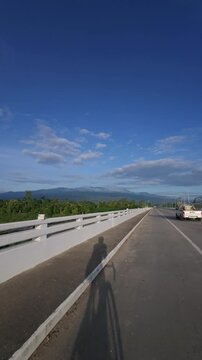 Shadow of a cyclist riding across a bridge toward a scenic mountain landscape beneath a clear blue sky, capturing outdoor travel, freedom, adventure and beautiful countryside scenery.