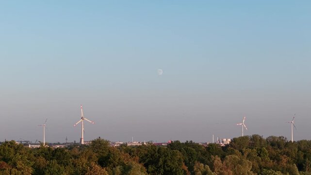 Wind turbines rise above dense woodland and distant industrial skyline beneath clear evening sky. Pale moon, warm sunlight, and hazy horizon create calm scenic renewable energy landscape in Europe