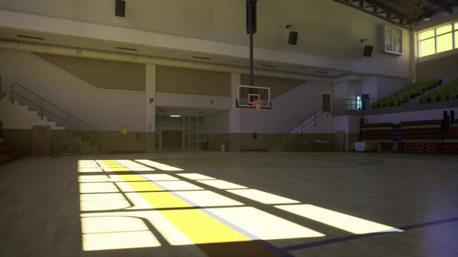 Golden hour empty gym basketball hoop silhouetted against bright windows, dramatic shadow patterns on polished hardwood, minimalist seating and high ceiling