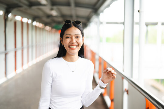 Happy and cheerful portrait of a young Thai woman smiling on a pedestrian skywalk bridge in Bangkok. She is wearing a simple yet stylish white long-sleeve tee, has sunglasses resting on her head, and 